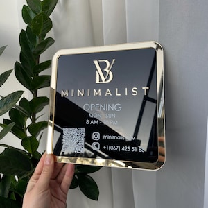 May include: A black and gold sign with the text "B MINIMALIST" and "OPENING MON-SUN 8 AM-10 PM". The sign also includes a QR code, Instagram handle, and phone number.