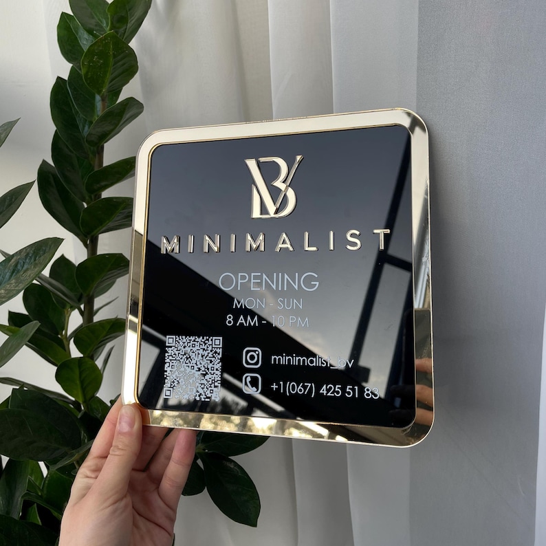 Op de afbeelding: Een zwart en gouden bord met de tekst "B MINIMALIST" en "OPENING MON-SUN 8 AM-10 PM". Het bord bevat ook een QR-code, een Instagram-handle en een telefoonnummer.