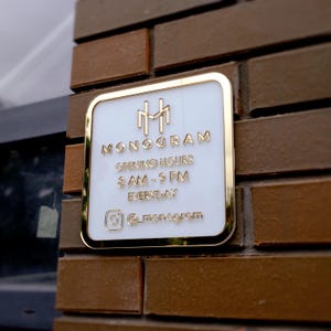 May include: A white rectangular sign with gold lettering and trim, fixed to a brick wall. The sign displays "MONOGRAM", opening hours from 8 AM to 9 PM daily, and an Instagram handle. The sign features a stylized logo.