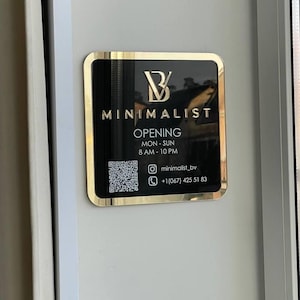 Op de afbeelding: Een gouden en zwart bord met de tekst "B MINIMALIST" en de woorden "OPENING MON-SUN 8 AM-10 PM" en contactgegevens, waaronder een QR-code, een Instagram-handvat en een telefoonnummer.