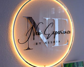 May include: Circular illuminated sign with the text "NE New Experience by Neshia". The sign has a silver metallic finish with a warm yellow glow around the edge. The sign is mounted on a light-colored wall.