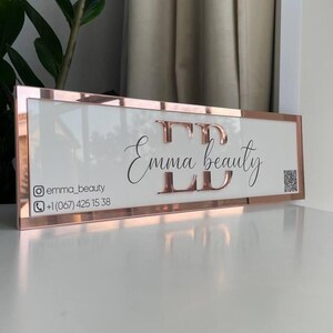 Op de afbeelding: Een wit bord met een ros&eacute;gouden frame en de tekst "Emma beauty" in zwart cursief schrift. Het bord bevat ook de social media handle "@emma_beauty" en een telefoonnummer.