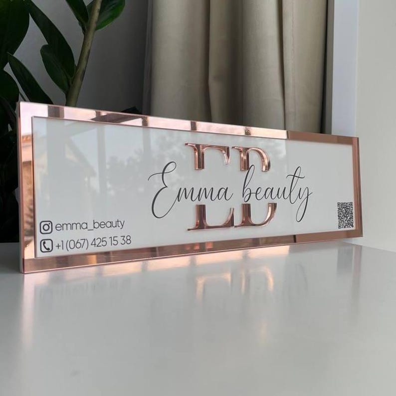 Op de afbeelding: Een wit bord met een ros&eacute;gouden frame en de tekst "Emma beauty" in zwart cursief schrift. Het bord bevat ook de social media handle "@emma_beauty" en een telefoonnummer.