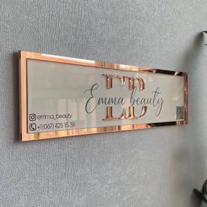 May include: Rose gold framed sign with the text "Emma beauty" and contact information for the business, including an Instagram handle and phone number.