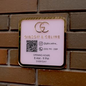 May include: A square, pink business sign with gold trim and lettering. The sign displays the business name "GIESON & CELINE", a QR code, social media handle, phone number, and opening hours: 8 AM - 9 PM everyday.