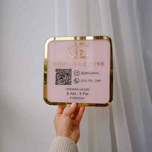 Op de afbeelding: Een vierkant, roze bord met gouden rand en een gouden logo met de tekst "GIBSON & CELINE". Het bord bevat een QR-code, Instagram-gebruikersnaam, telefoonnummer en openingstijden. Het bord wordt voor een wit gordijn gehouden.
