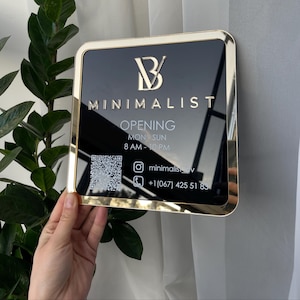 Könnte beinhalten: Ein quadratisches schwarzes Schild mit goldenem Rahmen, das das Wort "MINIMALIST" und die Öffnungszeiten zeigt. Das Schild enthält auch einen QR-Code, den Instagram-Handle "minimalisov" und eine Telefonnummer.