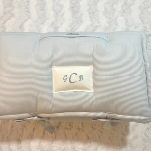 May include: A rectangular light blue pillow featuring a fine pinstripe pattern and a central cream-colored rectangular patch. The patch displays the embroidered monogram letters G, C, and B in a blue script font. The pillow has tufted details.