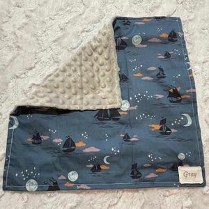 Puede incluir: Una manta para bebé azul y blanca con un respaldo de minky. La manta presenta un patrón de barcos navegando en un cielo nocturno estrellado. La manta tiene una etiqueta que dice "Gray".