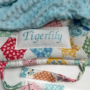May include: A patchwork quilt with a blue minky blanket on top. The quilt has a white label with the name "Tigerlily" embroidered in teal thread.