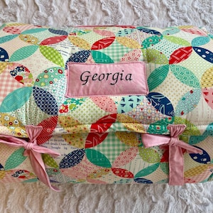 May include: A quilted, padded bag with a colorful patchwork design featuring various fabrics in red, blue, green, and pink. The bag has a pink label embroidered with the name "Georgia" and is tied with pink ribbons. The bag is approximately 12 inches long.