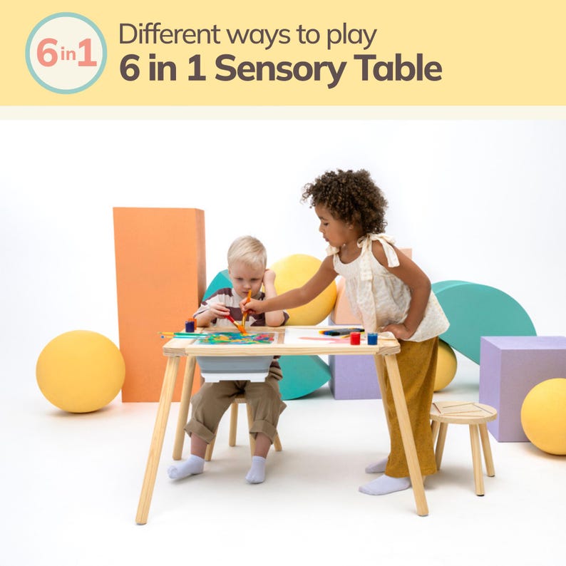 May include: Two children are playing at a wooden table with a white top. The table has a built-in tray and is labeled "6 in 1 Sensory Table". The table is surrounded by colorful shapes and a yellow ball.