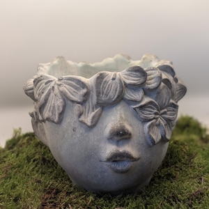 May include: A grey, face-shaped planter with a floral crown. The planter has a serene expression with closed eyes and a gentle smile. The detailed design features a textured surface and is set against a backdrop of green moss.