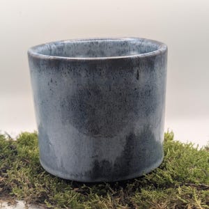May include: A cylindrical ceramic container with a dark blue glaze. The handmade pottery has a smooth, glossy finish and a slightly flared rim. The container is displayed on a bed of green moss, creating a natural aesthetic.