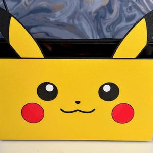 Puede incluir: Funda amarilla para la base de Nintendo Switch con temática de Pikachu. La funda presenta la cara de Pikachu con ojos negros, mejillas rojas y una boca sonriente. El diseño incluye las orejas de Pikachu.