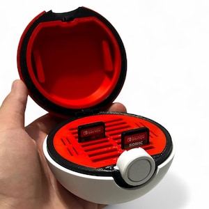 May include: A white, red, and black storage case for Nintendo Switch game cartridges. The case is spherical with a button closure and a red interior. Two game cartridges are visible inside the open case, one labeled "SONIC".