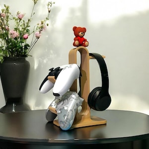 Könnte beinhalten: Ein Holzständer für Gaming-Zubehör mit zwei weißen und Camouflage-Gamecontrollern und einem schwarzen Kopfhörer. Ein kleiner, roter Teddybär sitzt oben auf dem Ständer. Der Ständer steht auf einem dunklen, runden Tisch.