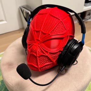 Può includere: Un supporto per cuffie a forma di maschera di Spider-Man rossa con cuffie nere e un microfono. Il supporto è strutturato con un motivo a ragnatela. Le cuffie hanno dettagli blu. Il microfono è nero.
