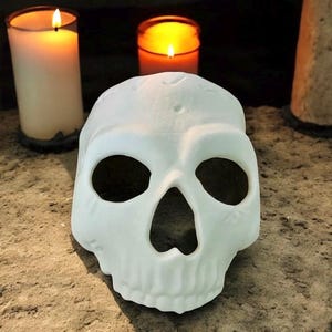May include: A white skull mask with large eye and nose openings rests on a textured surface. Two lit candles are in the background, creating a warm glow. The mask is a Halloween or Day of the Dead decoration.