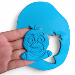 Pu&ograve; includere: Un tagliabiscotti in argilla blu a forma di volto di un personaggio dei cartoni animati, tenuto in mano. Il personaggio ha un ampio sorriso, grandi occhi e un ciuffo di capelli. Il tagliabiscotti &egrave; su una base di argilla blu pi&ugrave; grande.