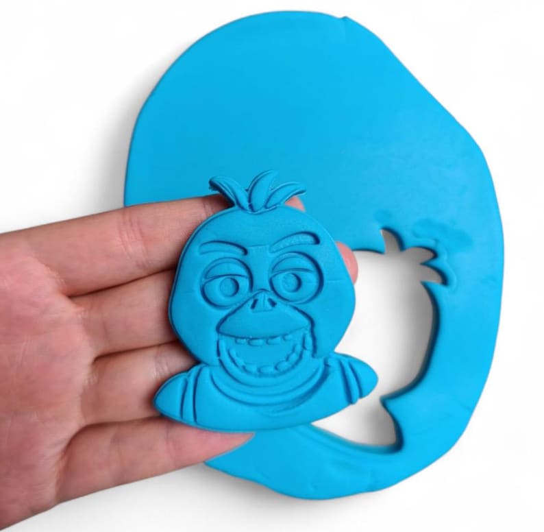 Pu&ograve; includere: Un tagliabiscotti in argilla blu a forma di volto di un personaggio dei cartoni animati, tenuto in mano. Il personaggio ha un ampio sorriso, grandi occhi e un ciuffo di capelli. Il tagliabiscotti &egrave; su una base di argilla blu pi&ugrave; grande.