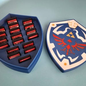 May include: A blue shield-shaped case and a tray holding Nintendo Switch game cartridges. The shield has a white border, a red bird emblem, and a gold crown design. The case is designed to hold the cartridges.