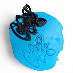 Pu&ograve; includere: Un tagliabiscotti in plastica nera a forma di testa di volpe dei cartoni animati, posto su una superficie blu con un'impronta dello stesso disegno. Il tagliabiscotti presenta dettagli come occhi, orecchie e muso.
