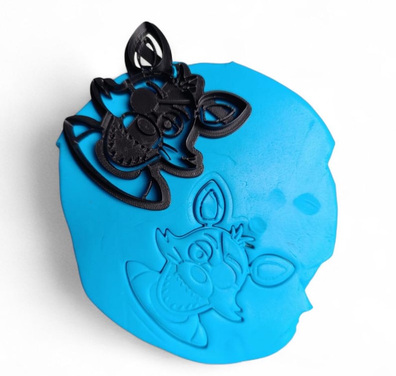 Pu&ograve; includere: Un tagliabiscotti in plastica nera a forma di testa di volpe dei cartoni animati, posto su una superficie blu con un'impronta dello stesso disegno. Il tagliabiscotti presenta dettagli come occhi, orecchie e muso.