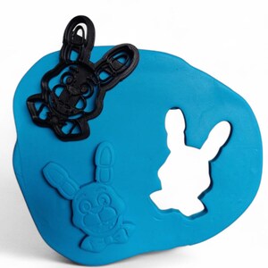 Pu&ograve; includere: Una base di plastilina blu con tre forme di coniglietto. Due tagliabiscotti neri a forma di coniglietto sono sopra, e un ritaglio a forma di coniglietto &egrave; nell'impasto. Le forme di coniglietto hanno dettagli, tra cui occhi, nasi e papillon.