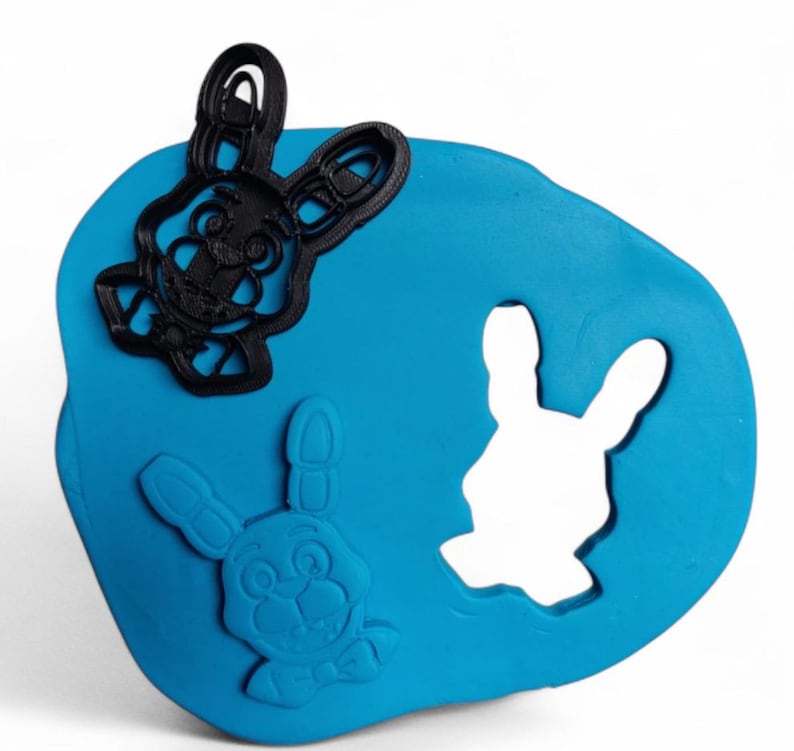 Pu&ograve; includere: Una base di plastilina blu con tre forme di coniglietto. Due tagliabiscotti neri a forma di coniglietto sono sopra, e un ritaglio a forma di coniglietto &egrave; nell'impasto. Le forme di coniglietto hanno dettagli, tra cui occhi, nasi e papillon.