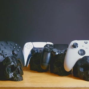 May include: A collection of black and white gaming controllers, including Nintendo Switch, PlayStation, and Xbox, resting on black skull-shaped stands. The controllers are arranged against a dark background, showcasing a gaming theme.