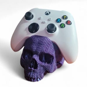 May include: A white Xbox controller rests atop a textured purple skull. The controller features black joysticks, colored buttons, and the Xbox logo. The skull has a detailed, woven appearance.