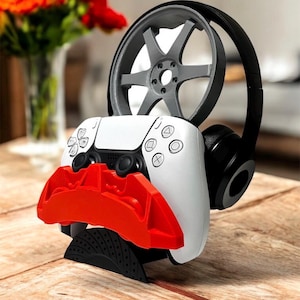 May include: A white and black gaming controller stand with a red accent, a grey wheel, and black headphones. The stand is designed to hold a gaming controller, headphones, and other accessories. The stand is on a wooden surface.