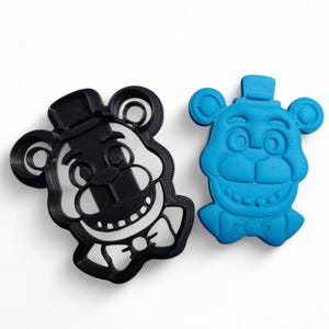 Pu&ograve; includere: Due formine per biscotti a forma di testa d'orso. Una &egrave; in bianco e nero, l'altra &egrave; blu. L'orso ha un cappello a cilindro, un papillon e un ampio sorriso. Le formine sono per fare biscotti a tema.