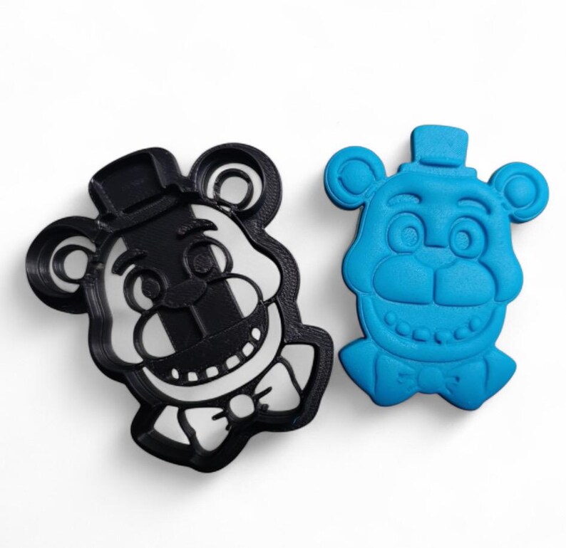 Pu&ograve; includere: Due formine per biscotti a forma di testa d'orso. Una &egrave; in bianco e nero, l'altra &egrave; blu. L'orso ha un cappello a cilindro, un papillon e un ampio sorriso. Le formine sono per fare biscotti a tema.