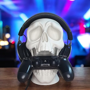 May include: A white skull-shaped headphone and controller stand. The stand holds a black gaming controller and black headphones. The background is a blurred mix of blue, purple, and green lights.