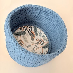 Könnte beinhalten: Ein blauer gehäkelter Korb mit einer weißen und grünen floralen Futter. Der Korb ist rund und hat eine gewebte Textur.