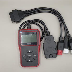 Pode incluir: Um scanner de diagnóstico OBD II preto e vermelho com vários cabos conectores. O scanner tem um ecrã e botões rotulados como "EXIT", "DTC" e "OK". Os cabos têm vários tipos de conectores, incluindo um preto grande e vários menores com detalhes coloridos.