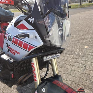 Könnte beinhalten: Nahaufnahme eines weißen und roten Yamaha Ténéré 700 Motorrads mit einem schwarzen Sturzbügel und einer klaren Windschutzscheibe. Das Motorrad hat ein schwarz-weißes "Ténéré 700" Logo und einen schwarzen "7" Aufkleber an der Seite.