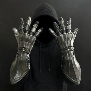 May include: A pair of articulated, dark gray metal gauntlets with riveted details. The armor-like gloves feature segmented fingers and a textured surface, suggesting a medieval or fantasy theme. The gauntlets are displayed against a dark background.