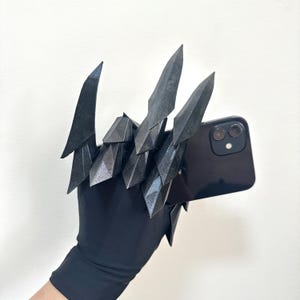 Puede incluir: Un guante negro con extensiones articuladas en forma de cristal puntiagudo unidas a los dedos. Las extensiones tienen una textura oscura, brillante y facetada. El guante sostiene un tel&eacute;fono inteligente negro con doble c&aacute;mara.