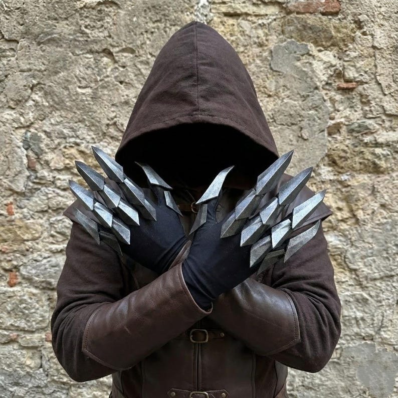 Puede incluir: Un par de guantes negros con accesorios met&aacute;licos plateados articulados en forma de garra en los dedos. Los guantes se llevan con una prenda de color marr&oacute;n oscuro con capucha que incluye brazaletes de cuero con hebillas met&aacute;licas.