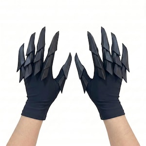 Puede incluir: Un par de guantes negros con estructuras articuladas en capas, similares a garras, que se extienden desde los dedos y el dorso de la mano. Los guantes est&aacute;n hechos de una tela oscura y lisa con accesorios r&iacute;gidos, puntiagudos y escamosos.