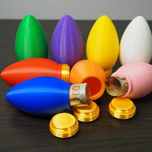 May include: A selection of brightly coloured, egg-shaped containers, some open to show rolled-up banknotes. The containers are in shades of green, purple, yellow, white, orange, red, blue, and pink, with gold-coloured screw-on lids.