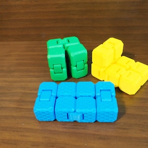 May include: Three plastic infinity cube fidget toys in bright solid colours. One cube is green, one is yellow, and one is blue. Each toy consists of multiple connected square blocks with visible hinges that allow the cubes to fold and unfold.