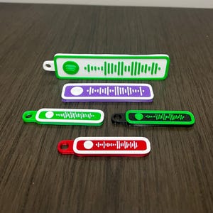 May include: Four different coloured keychains with a white background and a green barcode design. The keychains are green, purple, black and red.