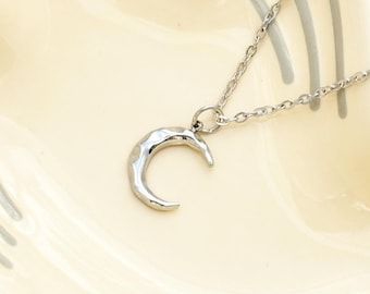 May include: A silver-colored necklace featuring a crescent moon pendant. The moon has a textured surface and hangs from a delicate chain. The necklace is displayed on a cream-colored surface with gray decorative lines.