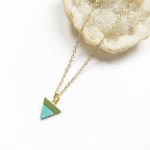 May include: A gold-colored necklace with a small turquoise and green triangle pendant. The pendant is a geometric shape, and the chain is delicate. The necklace is displayed on a white surface.