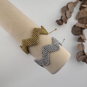 Könnte beinhalten: Zwei gewebte Armbänder mit einem ausgeprägten Zickzack- oder Chevron-Muster. Ein Armband ist aus metallischem Goldfaden gefertigt, das andere aus metallischem Silberfaden. Beide Artikel verfügen über verstellbare Kordelenden für das Handgelenk.