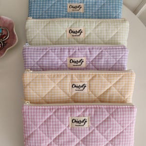 Può includere: Cinque astucci trapuntati in colori pastello: azzurro, verde, viola, giallo e rosa. Ogni astuccio presenta un motivo a quadretti e un'etichetta "Chiaragi Handmade". Gli astucci hanno una chiusura a cerniera.